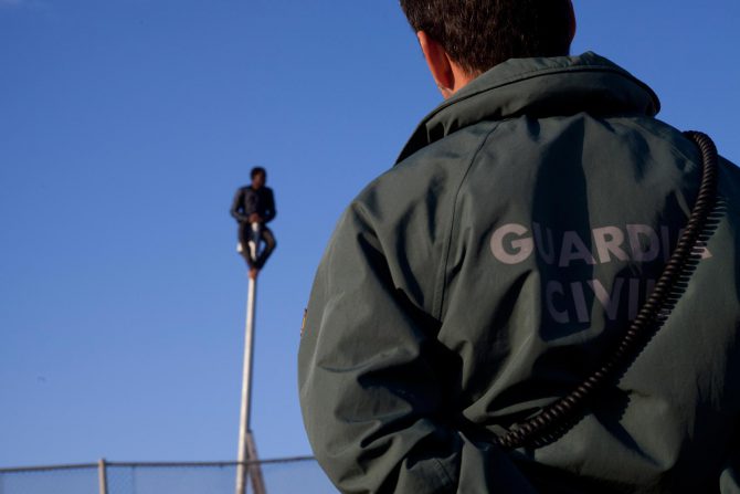 Un guardia civil observa a un subsahariano subido a un poste de la valla de Melilla.